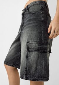 Bermuda cargo in denim con lavaggio nero. Le caratteristiche includono tasche laterali, una vestibilità leggermente ampia e cuciture a contrasto. La texture è resistente e ruvida.