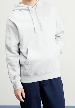 Osoba nosící světle šedou mikinu Nike s předním kapsou a stahovacími šňůrkami, kombinovanou s tmavě modrými kalhotami na jednobarevném pozadí.