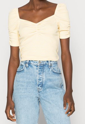 Żółto-biała bluzka w paski z krótkimi bufkowatymi rękawami i kwadratowym dekoltem, zestawiona z jasnoniebieskimi dżinsami z wysokim stanem.