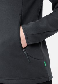 Giacca in pile con zip di colore grigio scuro, con una tasca laterale e coulisse elastica, caratterizzata da un'etichetta di marca verde vicino all'orlo per un tocco di stile.