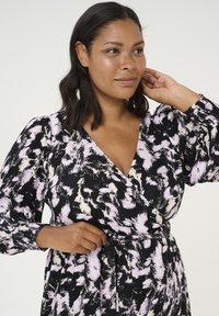 Vestido envolvente negro con un patrón abstracto en rosa y crema, mangas largas y detalle de cintura con lazo. Tela suave con un ligero brillo.