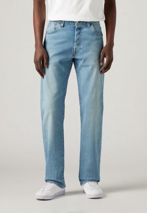 Homme portant un jean droit bleu clair et des baskets blanches, debout devant un fond uni clair.