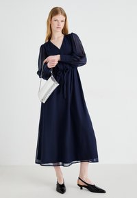 Navy blue, long-sleeve maxi dress with a V-neck, tied waist, and flowing fabric. Accessorized with a silver clutch and black pointed flats.