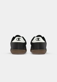 O pereche de adidași negri cu tab-uri albe la călcâi, având un logo "C" negru și talpa maro, văzuți din spate pe un fundal alb.