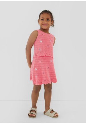 Jeune fille souriante, debout les mains derrière le dos, portant une robe rose sans manches en crochet avec des détails floraux et des sandales beiges.