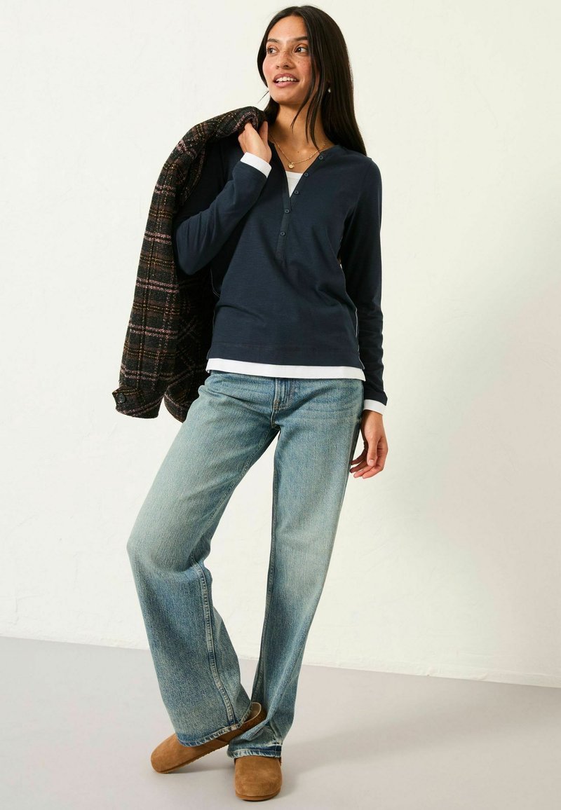 Marineblaues Langarmoberteil, weißes Unterhemd, hellblaue weit geschnittene Jeans, braune Clogs und eine dunkelgemusterte Jacke, die über die Schulter gelegt ist.