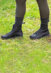 Piernas vestidas con leggings oscuros, calcetines gruesos grises y botas de tobillo negras con cordones, de pie sobre la hierba al aire libre.