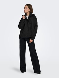 Black quilted jacket with a hood, featuring a front button closure and a relaxed fit. Worn over a light top and paired with dark wide-leg pants.