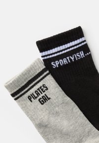 Két zokni: az egyik szürke, rajta a "PILATES GAL" felirattal fekete színben, a másik fekete, kék színű "SPORTYISH..." felirattal, mindkettőn vízszintes csíkok találhatók a tetején.