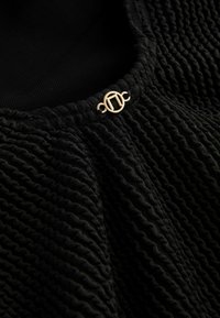 Gros plan sur un tissu noir texturé avec des vagues horizontales étroitement espacées et un petit fermoir rond doré présentant un design à double barre verticale.