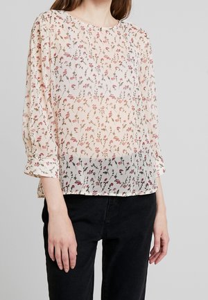 Nainen, joka käyttää läpikuultavaa, pitkähihamaista kermaista puseroa, jossa on pieni vaaleanpunainen ja ruskea kukkakuviointi, ja mustat housut.