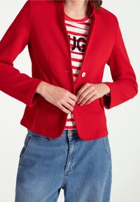 Red, textured blazer with a single button closure over a red and white striped shirt. Casual denim pants complete the outfit.