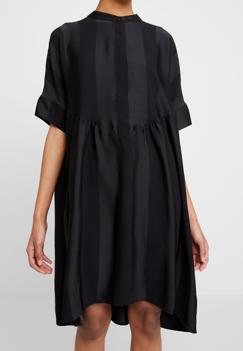 Kvinna i en löst sittande svart klänning med vertikala satinsränder, korta ärmar och samlad midjedetalj, stående mot en enfärgad bakgrund.