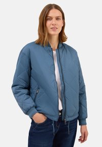 Femme avec des cheveux mi-longs portant une veste bomber matelassée bleue sur une chemise blanche et un jean noir, main dans la poche, regardant l'appareil photo.