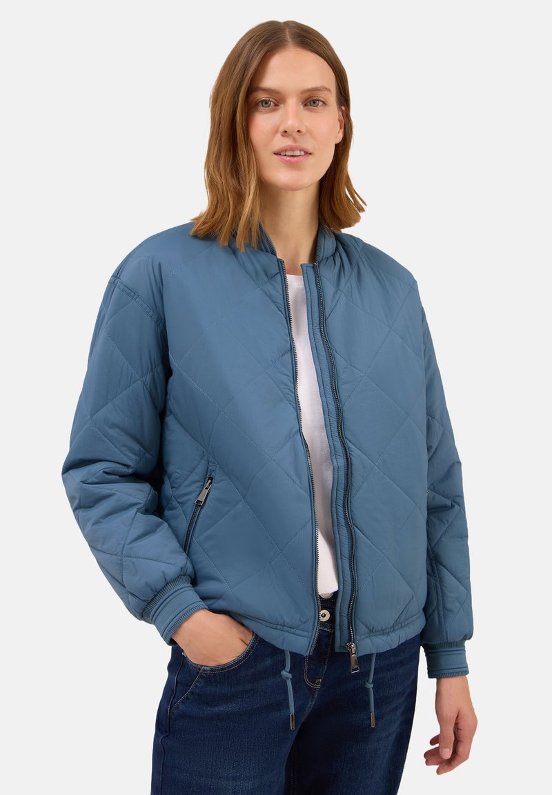Femme avec des cheveux mi-longs portant une veste bomber matelassée bleue sur une chemise blanche et un jean noir, main dans la poche, regardant l'appareil photo.