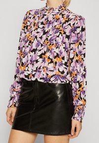 Blusa a maniche lunghe con motivo floreale in viola, arancione e nero, abbinata a una mini gonna in pelle nera con cucitura frontale e tasche.