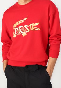 Sudadera roja con un logotipo gráfico dorado con motivo de cocodrilo y texto en negrita. Puños y cintura acanalados para mejorar el ajuste y la textura.