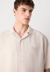 Kortermet, beige knappet skjorte laget av glatt stoff. Har en enkel krage, logo-lapp og subtil tekstur med runde knapper.