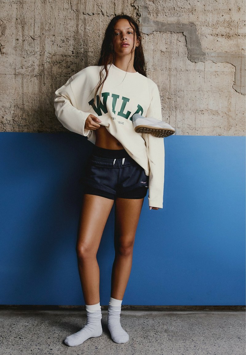 Jeune femme en chaussettes blanches, shorts noirs et sweat-shirt blanc oversize avec "WILD" se tient contre un mur bleu et en béton, tenant une basket.
