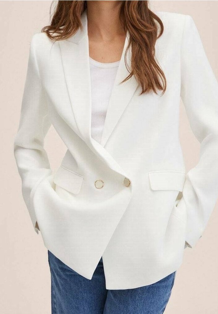 Kobieta w białym, dopasowanym żakiecie dwurzędowym z złotymi guzikami i niebieskich dżinsach, z rękami w kieszeniach, na neutralnym tle.