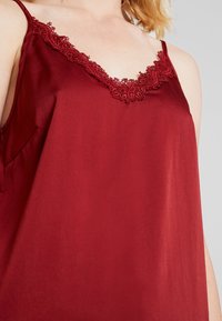 Rød satin cami med V-hals og intrikate blondedetaljer. Designet inkluderer tynde stropper og en glat, blank tekstur.