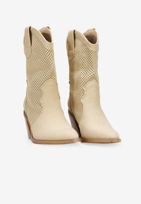 Botines de piel perforada beige con puntera afilada, acentos de diseño curvado y tacón apilado. La textura lisa realza el estilo moderno.