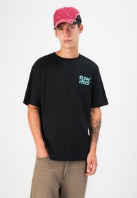 Sort kortærmet t-shirt med teksten "SLOW VIBES" i mintgrøn, parret med en lyserød kasket. Modellen har lysebrune bukser på med en lomme.