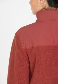 Rote Fleecejacke mit einem glatten oberen Bereich, hohem Kragen und strukturierter Körperpartie. Das Design ist praktisch mit einer körperbetonten Passform.