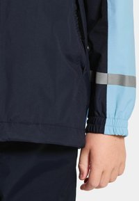 Ett barns hand bredvid en marinblå och ljusblå jackärm med en reflekterande grå rand och elastisk mudd, mot bakgrund av vit färg.