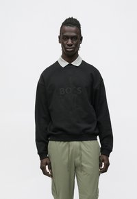 Μαύρο φούτερ με ανάγλυφο λογότυπο "BOSS", στρογγυλή λαιμόκοψη και γκρι πουκάμισο με γιακά από κάτω· σε συνδυασμό με πράσινο παντελόνι με κορδόνι.
