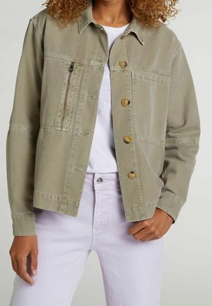 Femme portant une veste boutonnée couleur olive clair avec une poche poitrine zippée, chemise blanche et pantalon rose clair, debout avec la main dans la poche.