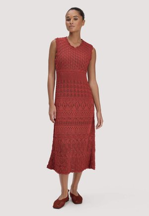 Vrouw in een roestkleurige mouwloze gebreide jurk met ingewikkelde patronen en bijpassende platte schoenen staand tegen een neutrale achtergrond.