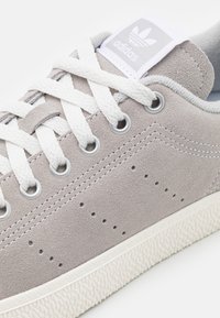 Sneaker in suede grigio con lacci bianchi e occhielli argentati. Presenta una suola in gomma testurizzata e una patch con il logo sulla linguetta.