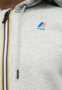 Φωτεινό γκρι φούτερ με πολύχρωμο φερμουάρ με ρίγες και μπλε και πορτοκαλί λογότυπο K-Way στο στήθος κοντά στον ώμο.