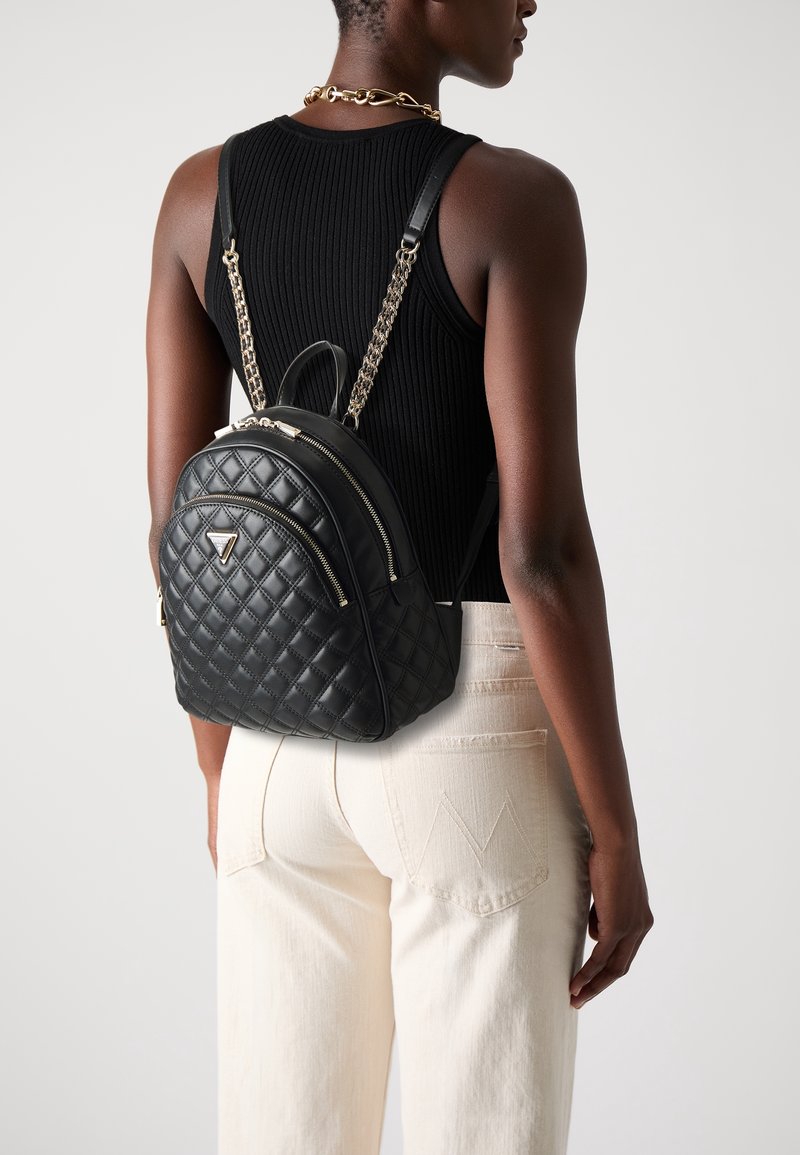 Femme portant un sac à dos en cuir matelassé noir avec des bretelles en chaîne dorée, un top noir sans manches, un collier en or et un pantalon blanc, vue de dos.