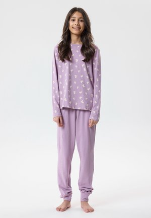 Młoda dziewczyna stojąca boso, ubrana w lawendową bluzkę z długim rękawem z białymi motylkami i pasujące spodnie na jasnym, jednolitym tle.