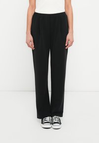 Μαύρο, άνετο παντελόνι από λείο υλικό με ελαστική μέση και πιέτες μπροστά, συνδυασμένο με μαύρα και λευκά αθλητικά παπούτσια.