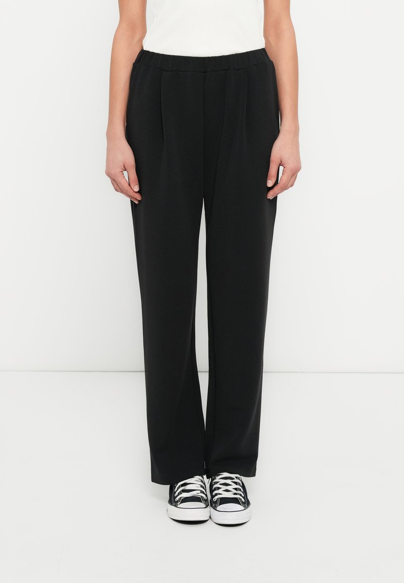 Μαύρο, άνετο παντελόνι από λείο υλικό με ελαστική μέση και πιέτες μπροστά, συνδυασμένο με μαύρα και λευκά αθλητικά παπούτσια.