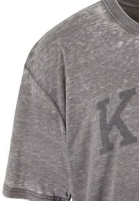 Pilka, trumpa rankovė t-shirt su minkštu, tekstūriniu paviršiumi ir dideliu neryškiu juodu raide "K" priekyje. Kasdienis kirpimas, apvalus iškirpimas.