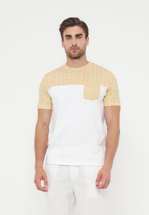 Homme debout, face à l'avant, portant un t-shirt blanc avec une partie supérieure jaune à motifs et une poche poitrine, associé à un pantalon blanc.