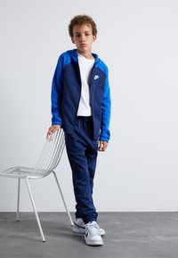 Nike trainingspak in blauwtinten bestaat uit een zip-up jack met capuchon en elastische manchetten, gecombineerd met bijpassende broeken. Witte sneakers maken de look compleet.