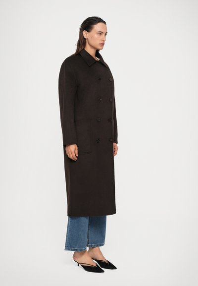 Kvinde står iført en lang, mørkebrun dobbeltbrystet frakke, beskårede blå jeans og sorte kitten heel mules på en hvid baggrund.