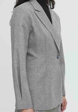 Kvinde iført en skræddersyet grå blazer med en enkelt knap over en sort turtleneck, vist fra skuldre til hofter på en ensfarvet baggrund.