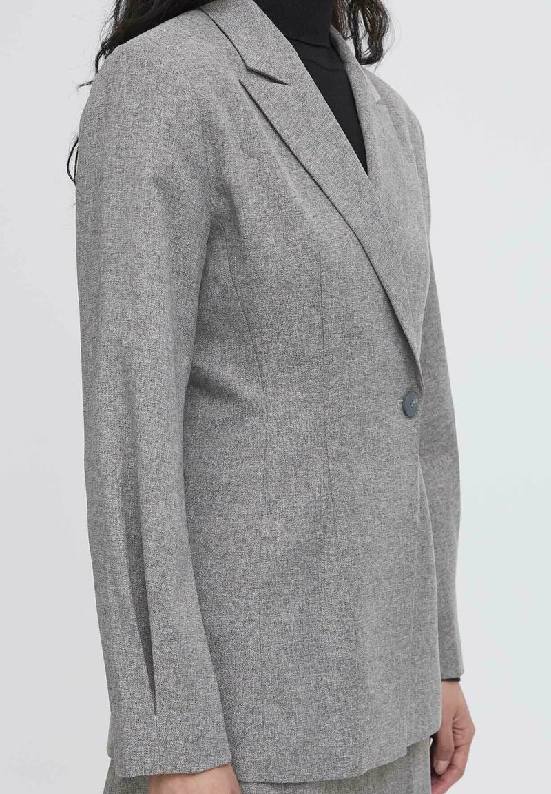Donna che indossa un blazer grigio su misura con un bottone sopra un dolcevita nero, mostrata dalle spalle ai fianchi su uno sfondo neutro.