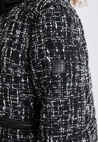 Black and white tweed jacket with a textured pattern, featuring a black logo patch and a zippered pocket detail.
