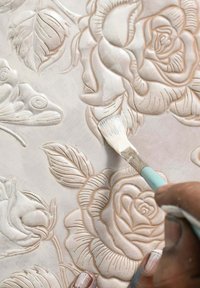 Relief-Dekorationswandpaneel mit erhabenen Mustern von Rosen und Blättern. Ein Pinsel trägt weiße Farbe auf und hebt Details und Texturen hervor.