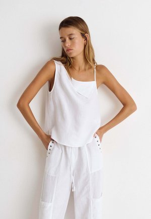 Mujer joven con cabello largo que lleva una camiseta blanca asimétrica sin mangas y pantalones blancos holgados con las manos en los bolsillos sobre fondo blanco.