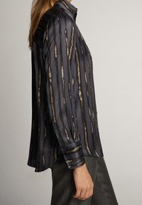Femme portant une chemise rayée noire et marron à manches longues boutonnée, associée à un pantalon gris foncé, vue de profil.