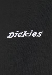 Bílá vyšívaná logo "Dickies" na černé texturované látce, pravděpodobně součást oděvu.