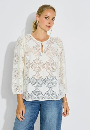 Mujer rubia con una blusa blanca de encaje con mangas abullonadas y vaqueros azules, de pie frente a un fondo claro y liso.